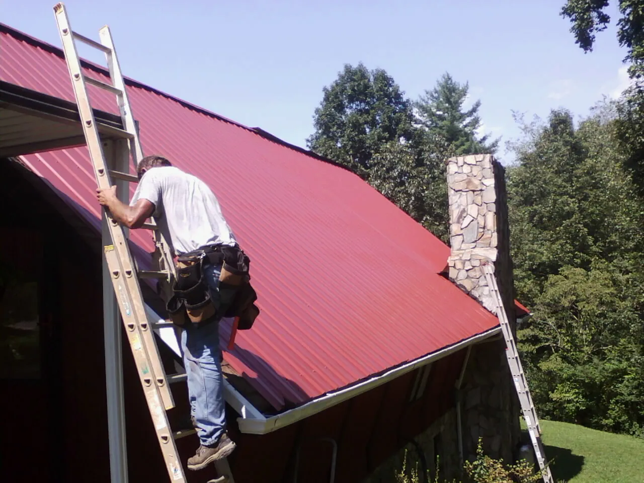 Roofer on a ladder beside a red metal roof and stone chimney — Soffit & Fascia Repair by Roofing Brothers in Sturgeon Bay